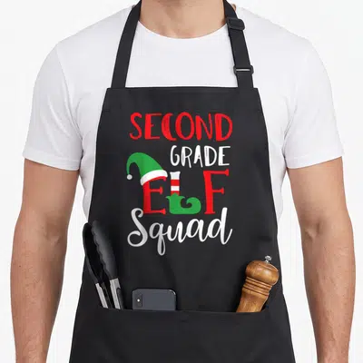 Second christmas elf apron - Stacked SECOND GRADE ELF Squad text with cartoon elf hat and shoes in red, white, and green — shown on a black cotton canvas apron against a white background
