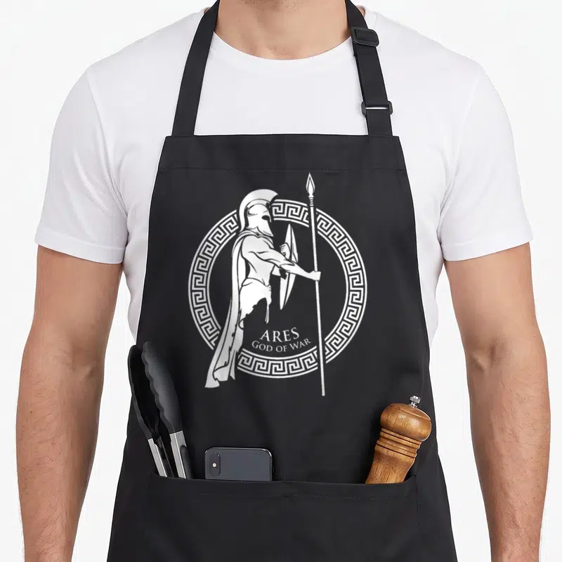 History greek aprons - White line art of helmeted warrior Ares with shield spear and Greek key border — shown on a black cotton canvas apron against a white background
