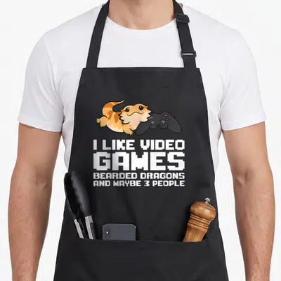 Maybe video game apron - Orange cartoon bearded dragon holding black controller with white pixel text below — shown on a black cotton canvas apron against a white background