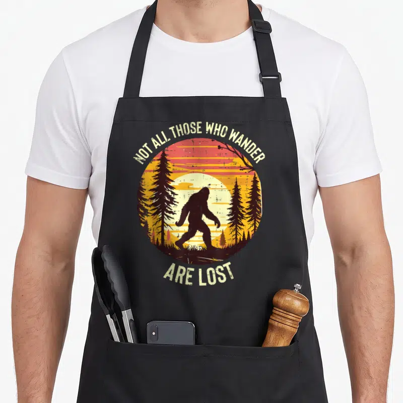Wander lost bigfoot apron - Retro circular sunset with black Bigfoot silhouette walking through pines and arching text — shown on a black cotton canvas apron against a white background