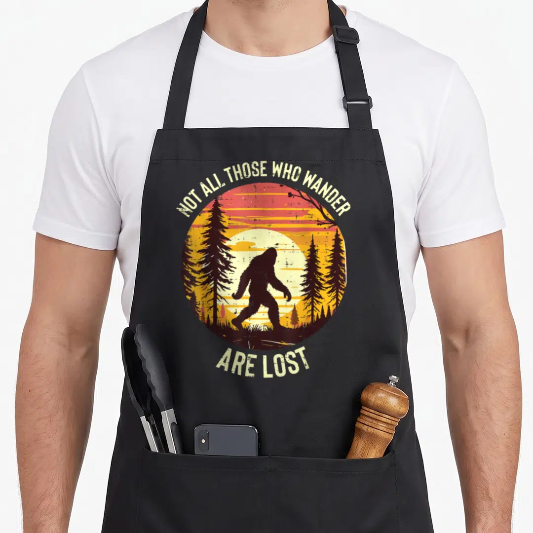 Wander lost bigfoot apron - Retro circular sunset with black Bigfoot silhouette walking through pines and arching text — shown on a black cotton canvas apron against a white background