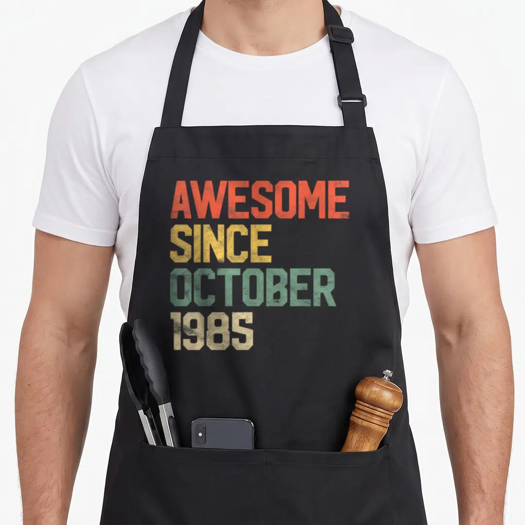 October 35th birthday apron - Stacked AWESOME SINCE OCTOBER 1985 text in distressed orange, yellow, green, and beige — shown on a black cotton canvas apron against a white background