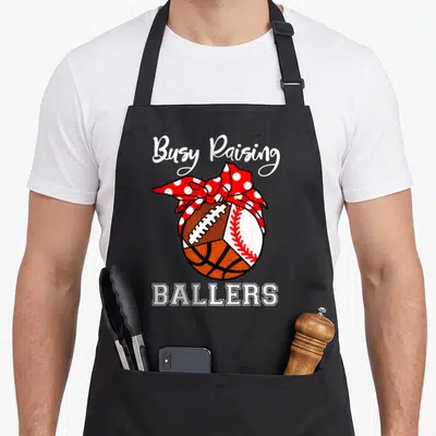 Football busy apron - Composite ball with football basketball baseball and red polka dot bow text Busy Raising Ballers — shown on a black cotton canvas apron against a white background