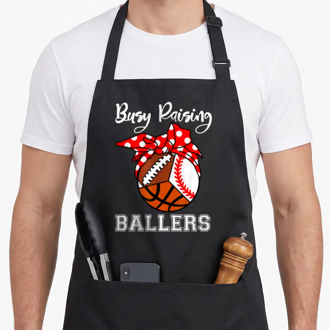 Football busy apron - Composite ball with football basketball baseball and red polka dot bow text Busy Raising Ballers — shown on a black cotton canvas apron against a white background