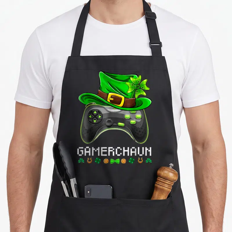 Video st patrick day aprons - Black game controller with green leprechaun hat and pixel text GAMERCHAUN — shown on a black cotton canvas apron against a white background