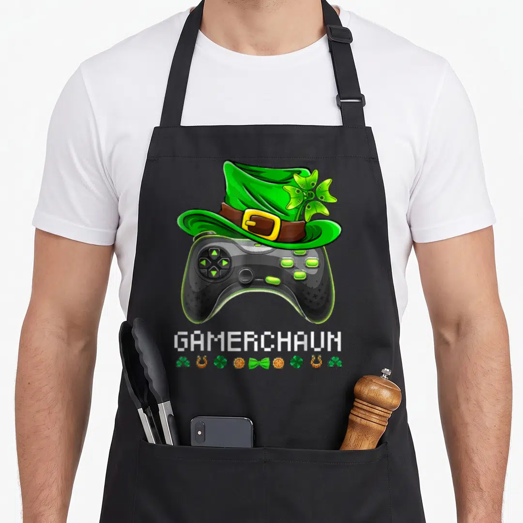 Video st patrick day aprons - Black game controller with green leprechaun hat and pixel text GAMERCHAUN — shown on a black cotton canvas apron against a white background