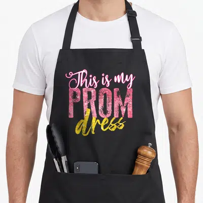 School prom apron - Stacked pink and yellow glitter text reading This is my PROM dress — shown on a black cotton canvas apron against a white background