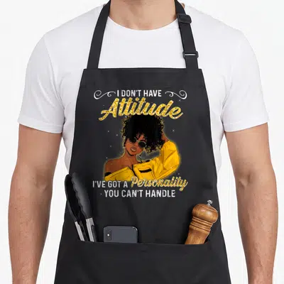 Melanin queen african american aprons - Cartoon woman in yellow jacket with gold glitter text I DON'T HAVE Attitude — shown on a black cotton canvas apron against a white background