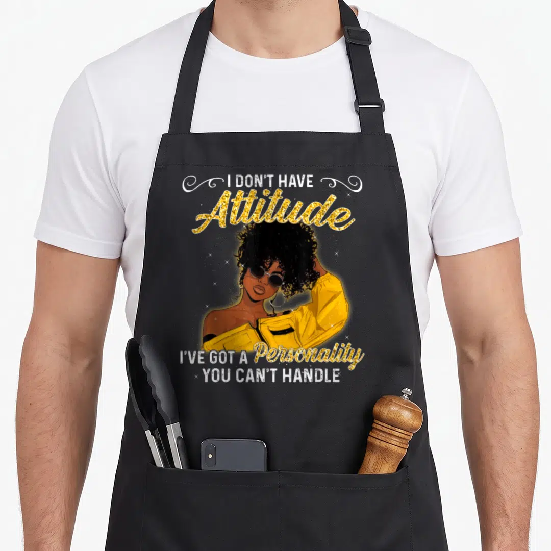 Melanin queen african american aprons - Cartoon woman in yellow jacket with gold glitter text I DON'T HAVE Attitude — shown on a black cotton canvas apron against a white background
