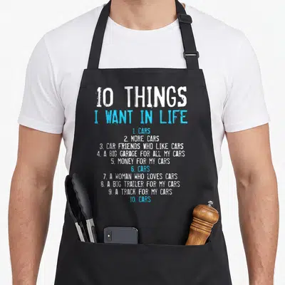 Retro car garage apron - White and blue typography listing 10 things wanting cars garage trailer track money woman — shown on a black cotton canvas apron against a white background