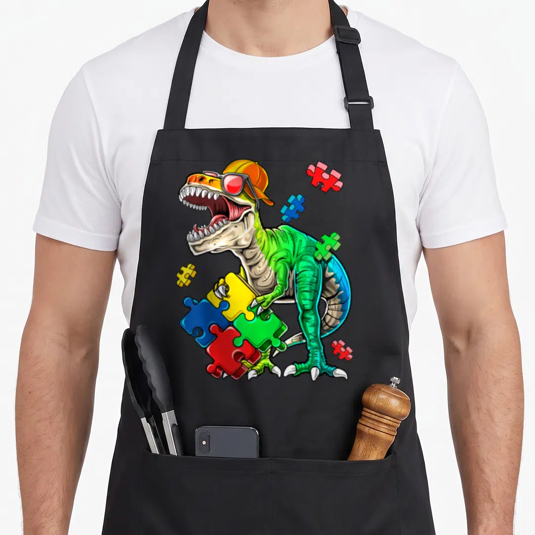 Rex autism dinosaur apron - Cartoon T-Rex with orange cap and sunglasses holding colorful puzzle pieces — shown on a black cotton canvas apron against a white background