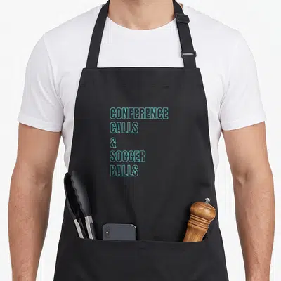 Playful game aprons - Cyan outline text reading CONFERENCE CALLS & SOCCER BALLS stacked vertically — shown on a black cotton canvas apron against a white background