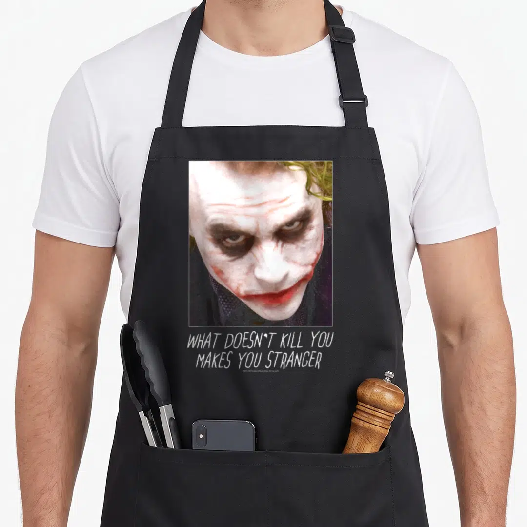Dark fools apron - Joker face photo above white text WHAT DOESN'T KILL YOU MAKES YOU STRANGER on black background — shown on a black cotton canvas apron against a white background