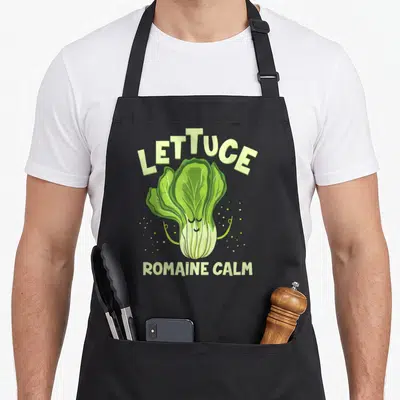 Romaine vegan apron - Cartoon green lettuce with closed eyes and 'Lettuce Romaine Calm' text surrounded by small dots — shown on a black cotton canvas apron against a white background