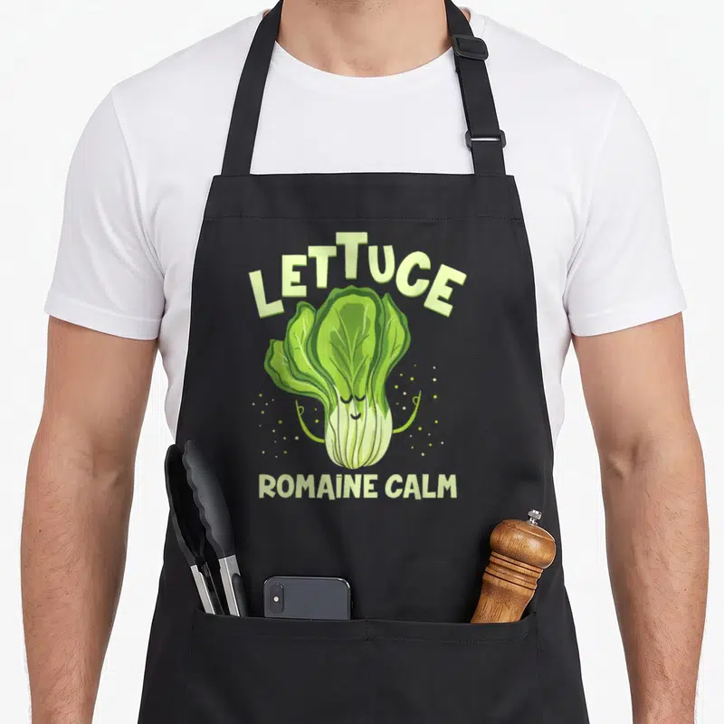 Romaine vegan apron - Cartoon green lettuce with closed eyes and 'Lettuce Romaine Calm' text surrounded by small dots — shown on a black cotton canvas apron against a white background