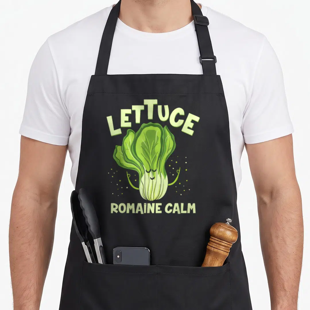 Romaine vegan apron - Cartoon green lettuce with closed eyes and 'Lettuce Romaine Calm' text surrounded by small dots — shown on a black cotton canvas apron against a white background