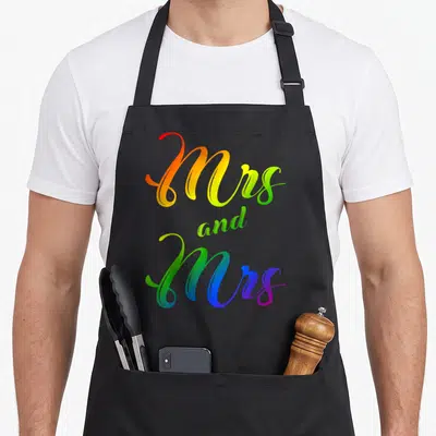 Lesbian mrs apron - Rainbow gradient script text reading Mrs and Mrs stacked vertically — shown on a black cotton canvas apron against a white background