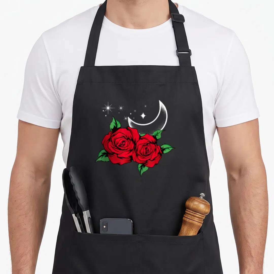 Night gardener apron - Two red roses with green leaves below a white crescent moon and scattered stars — shown on a black cotton canvas apron against a white background
