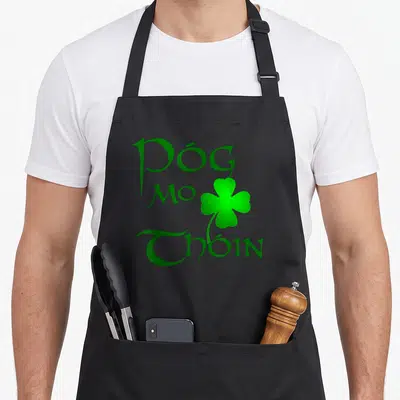 Gaelic irish aprons - Green Póg Mo Thóin text and shamrock on black background — shown on a black cotton canvas apron against a white background