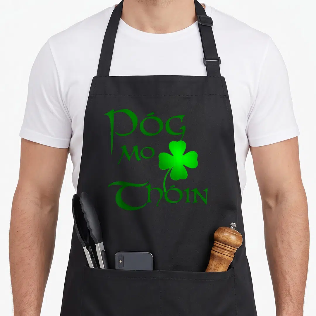 Gaelic irish aprons - Green Póg Mo Thóin text and shamrock on black background — shown on a black cotton canvas apron against a white background