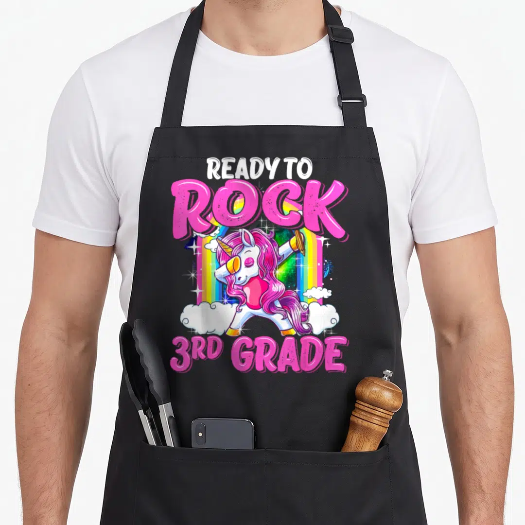Rock unicorn apron - Dabbing unicorn with pencil, rainbow, clouds, READY TO ROCK 3RD GRADE text in pink white yellow — shown on a black cotton canvas apron against a white background