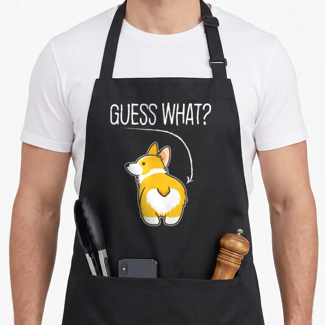 Guess welsh apron - Cartoon corgi rear view with white arrow and GUESS WHAT text — shown on a black cotton canvas apron against a white background