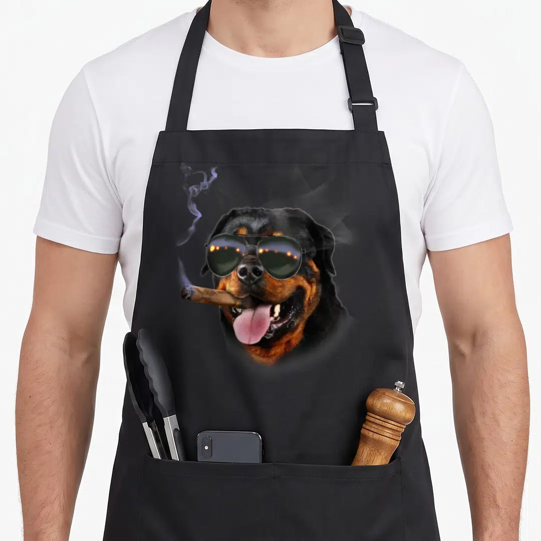 Rottweiler stylish apron - Rottweiler head wearing aviator sunglasses and holding a smoking cigar in photo style — shown on a black cotton canvas apron against a white background
