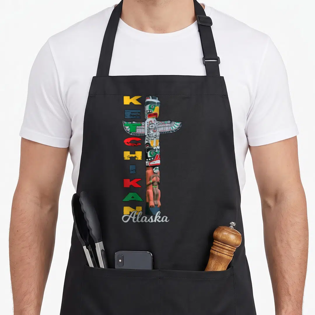 Alaska ketchikan apron - Vertical totem pole with stacked KETCHIKAN letters and script Alaska label in yellow red green — shown on a black cotton canvas apron against a white background