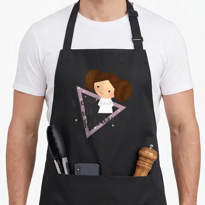 Rule kawaii apron - Cartoon girl with brown buns in white dress above purple triangle and Moms Rule The Galaxy text — shown on a black cotton canvas apron against a white background