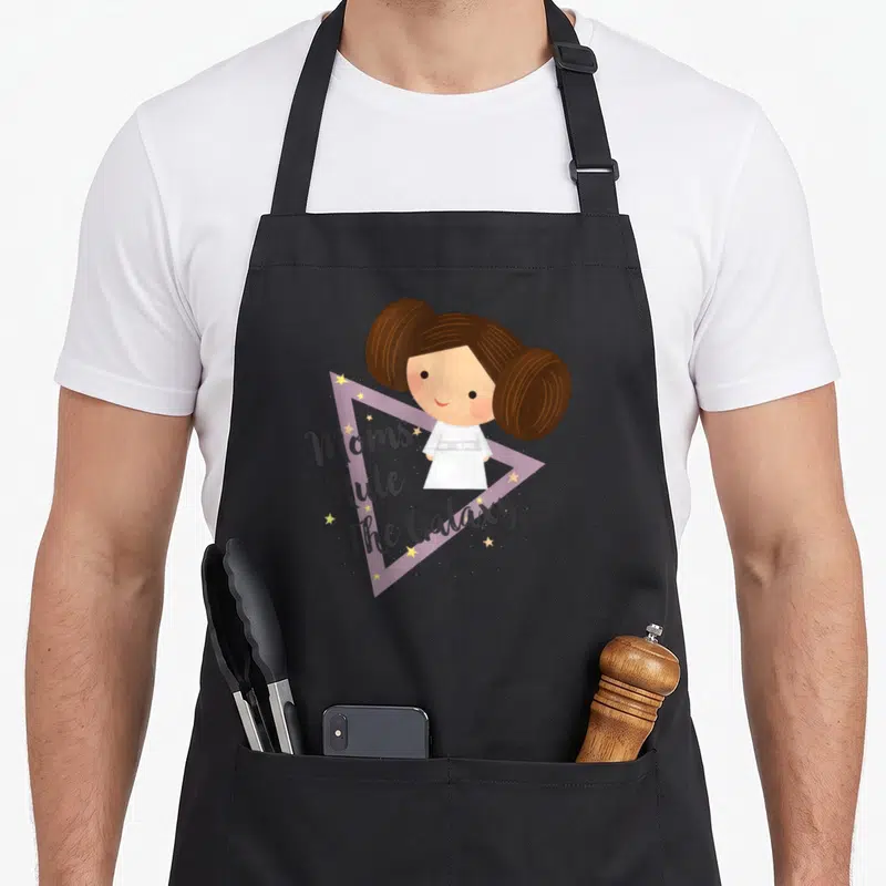 Rule kawaii apron - Cartoon girl with brown buns in white dress above purple triangle and Moms Rule The Galaxy text — shown on a black cotton canvas apron against a white background