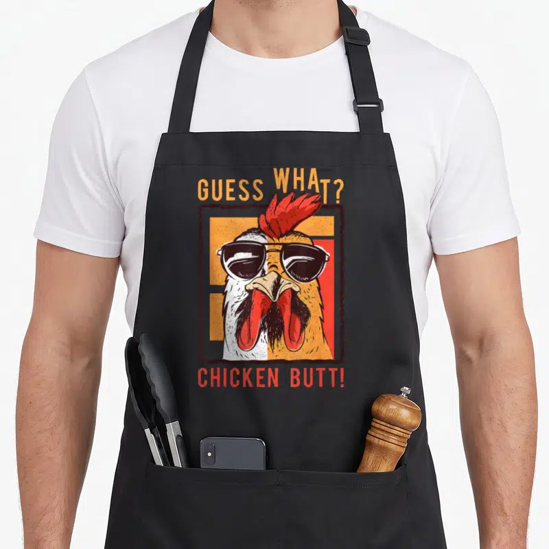 Sibling chicken apron - Cartoon rooster with sunglasses and Guess What Chicken Butt text — shown on a black cotton canvas apron against a white background