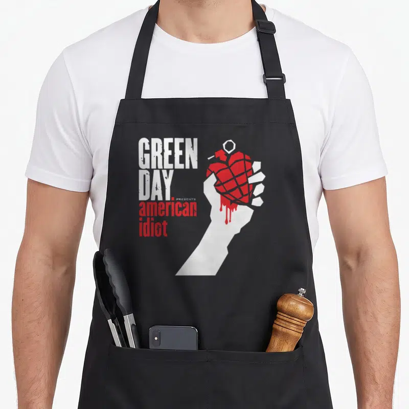 Idiot comfort apron - White hand holding red grenade next to stacked Green Day American Idiot text — shown on a black cotton canvas apron against a white background