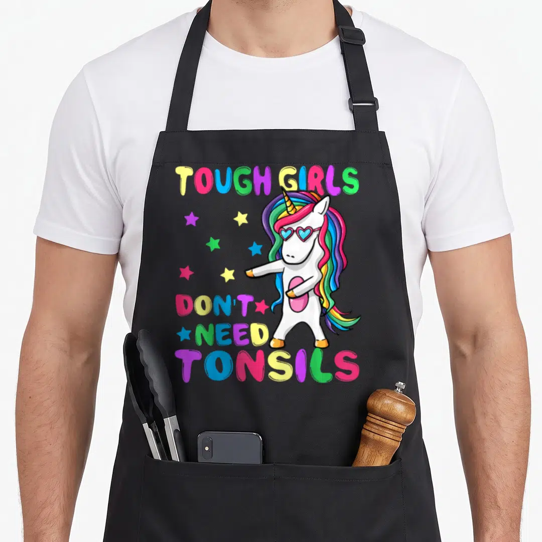 Tough wellness apron - Dabbing unicorn with rainbow mane and heart sunglasses surrounded by multi-colored stars and text — shown on a black cotton canvas apron against a white background