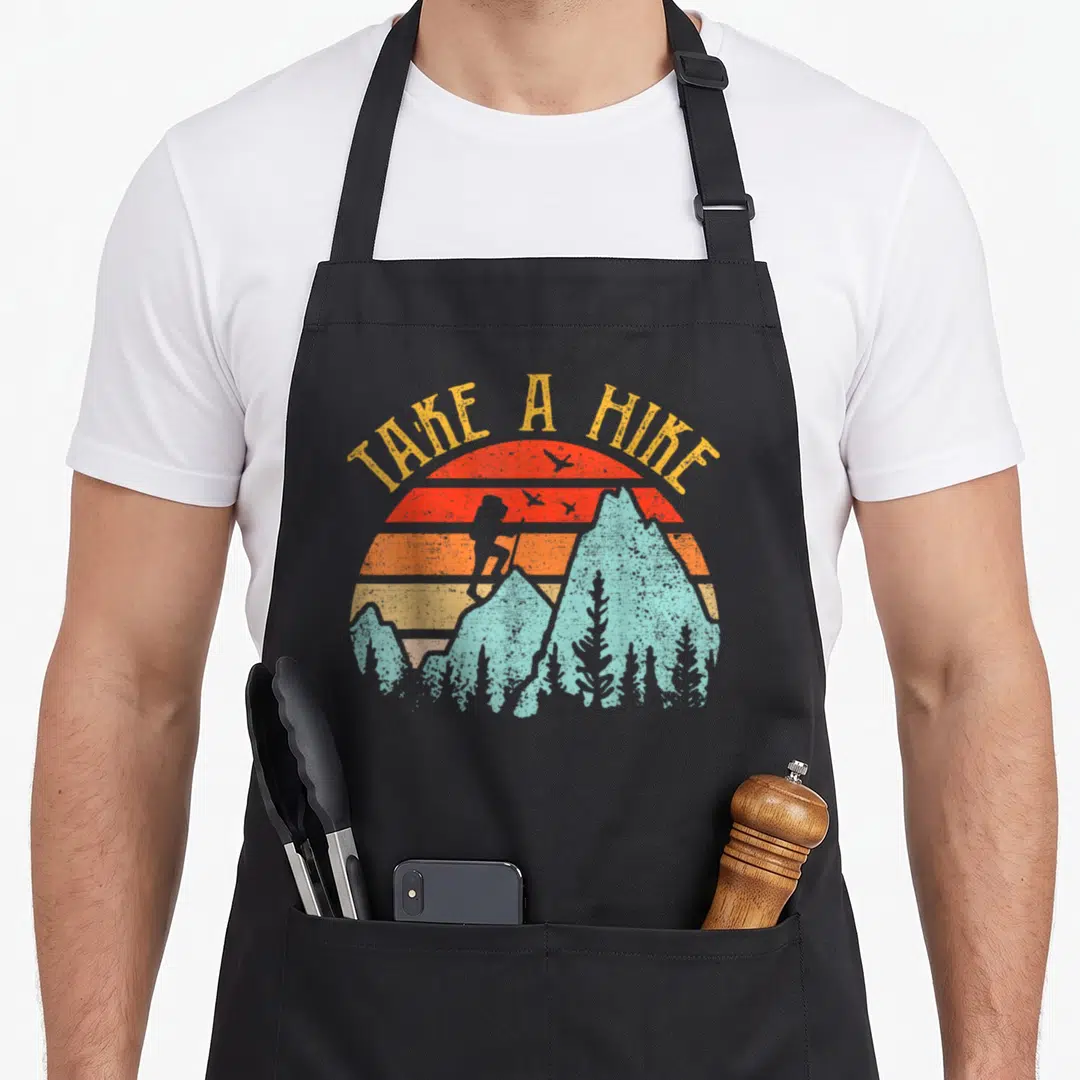 Hiking hike camping apron - Retro mountain landscape with arched TAKE A HIKE text and striped sun containing a hiker — shown on a black cotton canvas apron against a white background