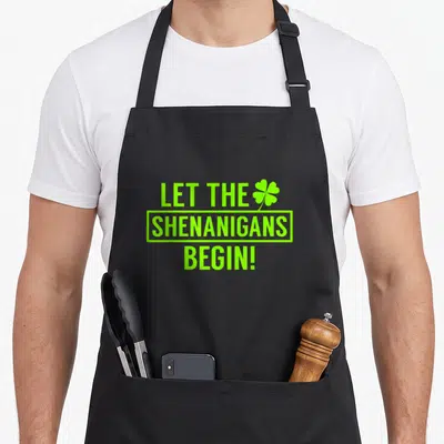 Shenanigan irish stout apron - Lime green stacked text reading LET THE SHENANIGANS BEGIN with a four-leaf clover icon — shown on a black cotton canvas apron against a white background