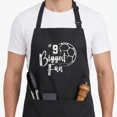 Biggest family aprons - White distressed #9's Biggest Fan text and soccer ball outline — shown on a black cotton canvas apron against a white background