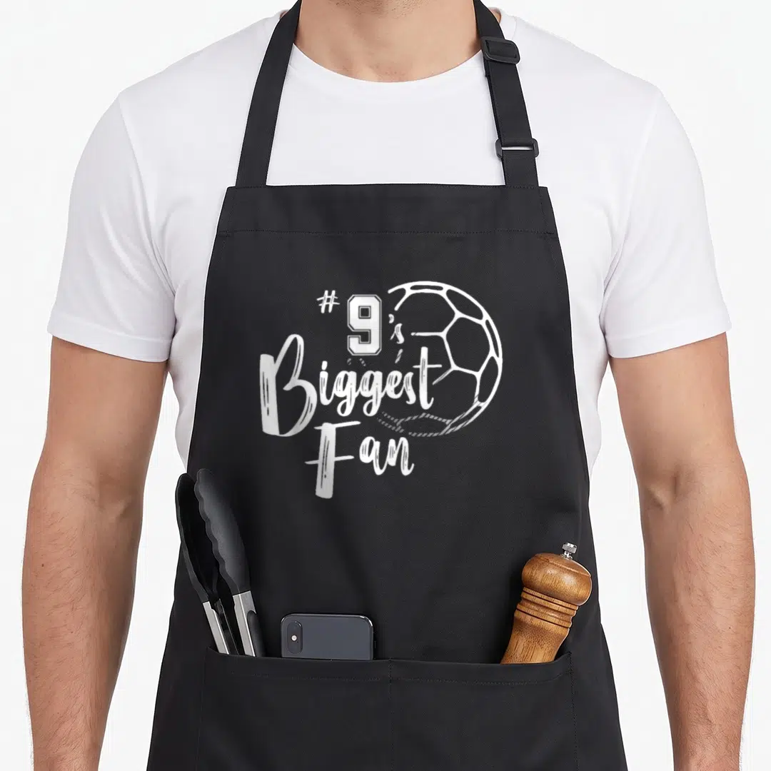 Biggest family aprons - White distressed #9's Biggest Fan text and soccer ball outline — shown on a black cotton canvas apron against a white background