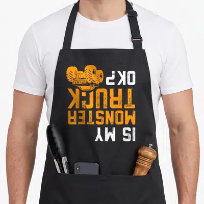 Offroad truck apron - Upside down orange monster truck and Is My Monster Truck OK text — shown on a black cotton canvas apron against a white background