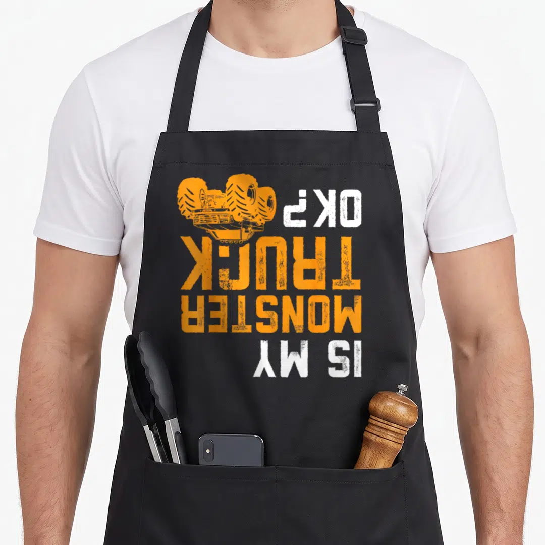 Offroad truck apron - Upside down orange monster truck and Is My Monster Truck OK text — shown on a black cotton canvas apron against a white background