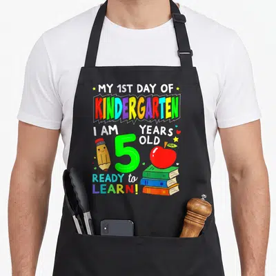 1st day kindergarten apron - Cartoon pencil, green number 5, stacked books, red apple, and stars scattered around — shown on a black cotton canvas apron against a white background