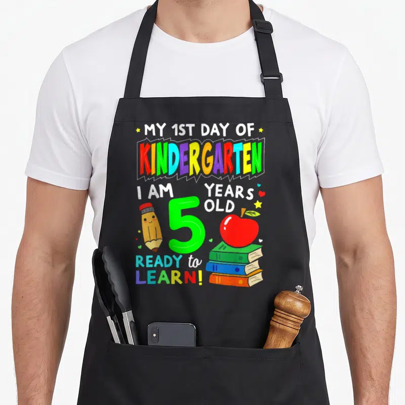 1st day kindergarten apron - Cartoon pencil, green number 5, stacked books, red apple, and stars scattered around — shown on a black cotton canvas apron against a white background