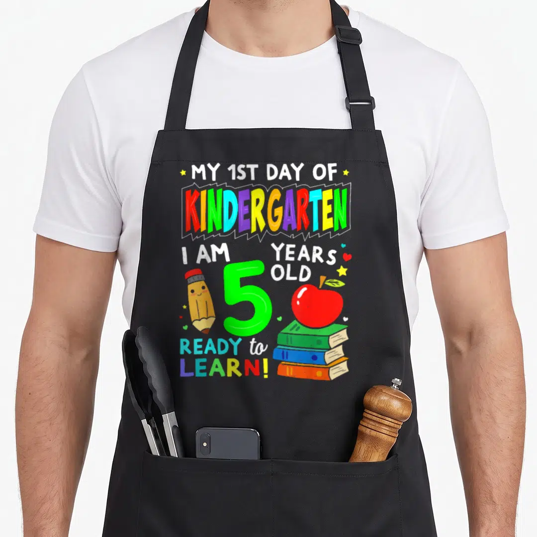 1st day kindergarten apron - Cartoon pencil, green number 5, stacked books, red apple, and stars scattered around — shown on a black cotton canvas apron against a white background