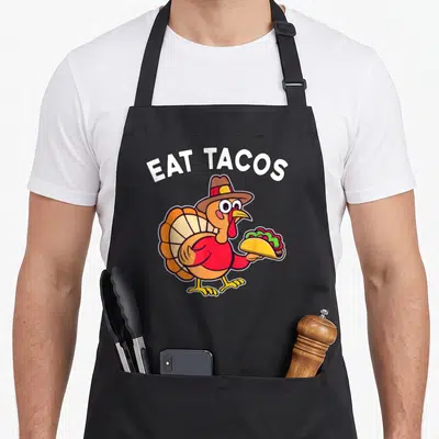 Eat turkey apron - Cartoon turkey in a hat holding a taco under arched white EAT TACOS text — shown on a black cotton canvas apron against a white background