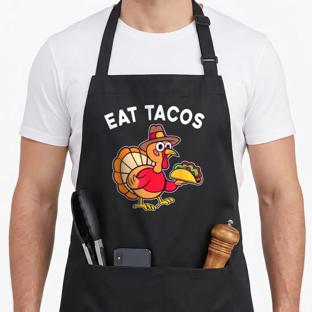 Eat turkey apron - Cartoon turkey in a hat holding a taco under arched white EAT TACOS text — shown on a black cotton canvas apron against a white background