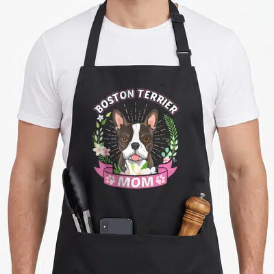 Terrier mother's day aprons - Cartoon Boston Terrier head with flowers and MOM ribbon in brown, pink, white, and green — shown on a black cotton canvas apron against a white background