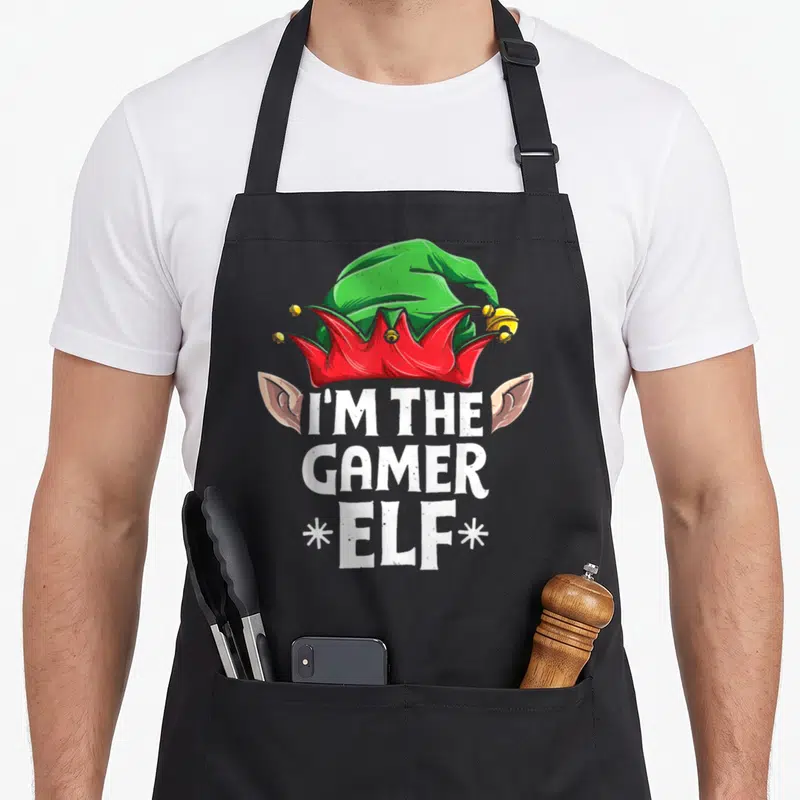 Family pyjamas christmas elf apron - Green elf hat with red trim, white I'M THE GAMER ELF text, two ears, two — shown on a black cotton canvas apron against a white background
