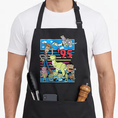 Distressed colorful apron - Toy Story characters clustered under red '95 text against blue sky and colorful horizontal — shown on a black cotton canvas apron against a white background