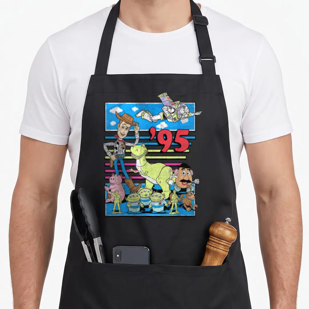 Distressed colorful apron - Toy Story characters clustered under red '95 text against blue sky and colorful horizontal — shown on a black cotton canvas apron against a white background