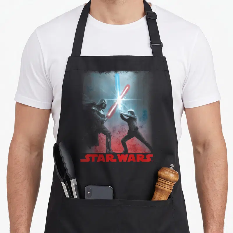 Battle general apron - Darth Vader and Luke Skywalker clash with red and blue lightsabers beneath STAR WARS text — shown on a black cotton canvas apron against a white background