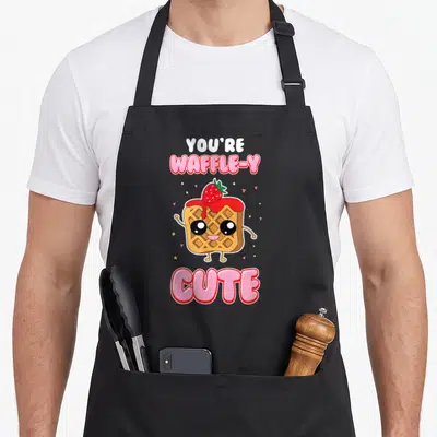 Awfully waffles apron - Cartoon waffle with syrup strawberry and text Youre Waffle-y Cute surrounded by hearts — shown on a black cotton canvas apron against a white background